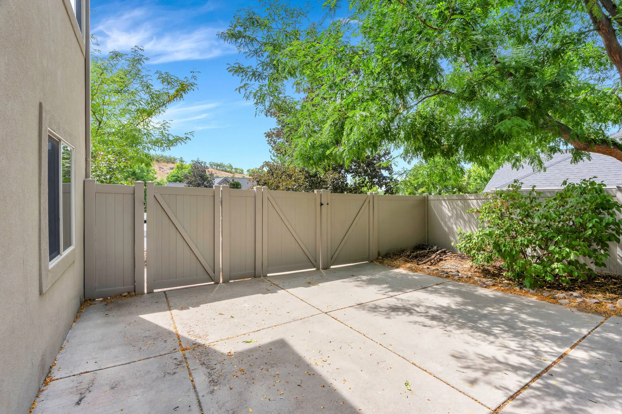 Paved patio area featuring a privacy fence, mature trees, and exterior window