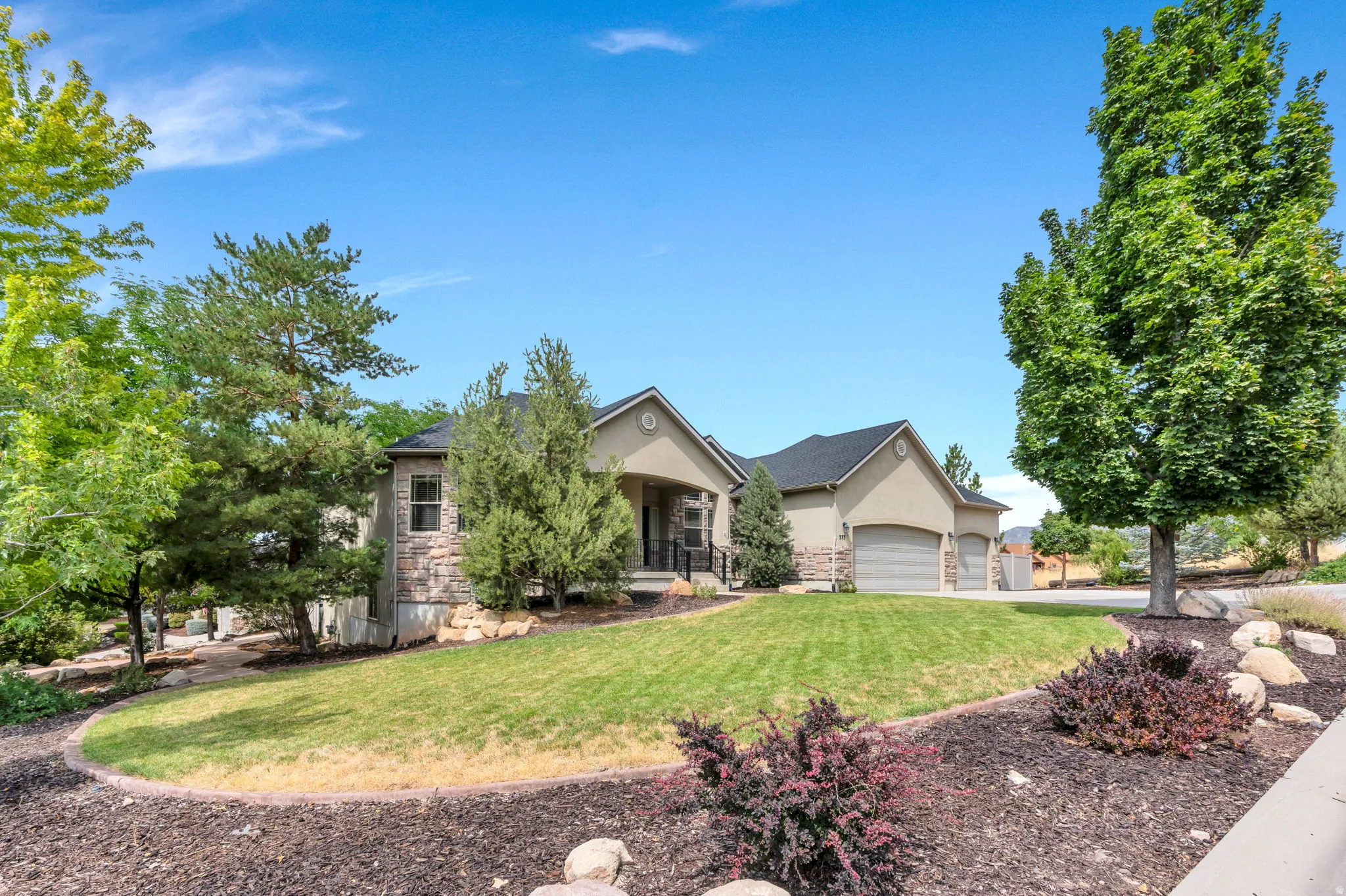 Stucco and stone exterior, covered entry, attached garages, established landscaping, and a sloped lawn