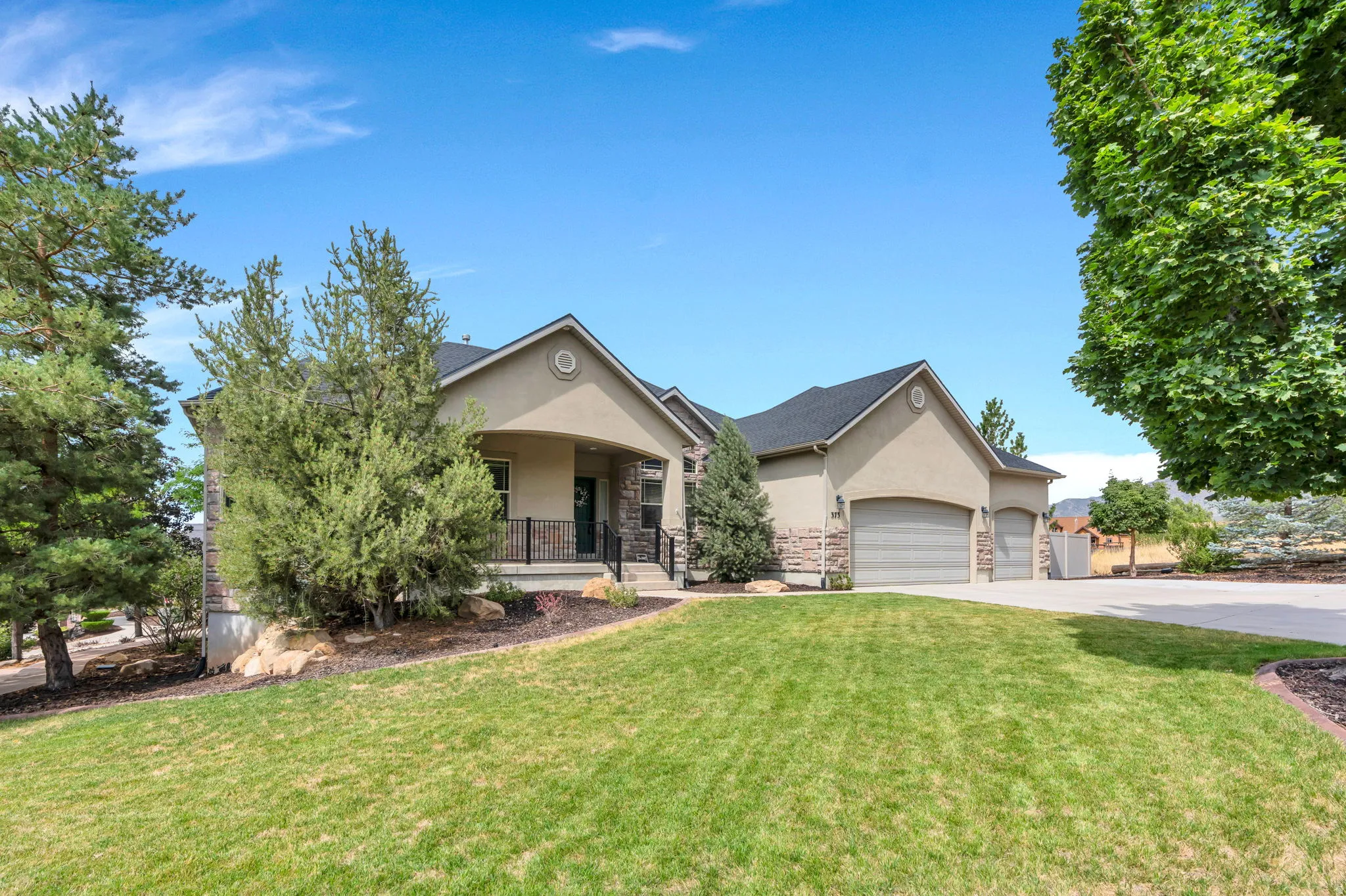 Stucco exterior with stone accents, covered front porch with iron railing, attached multiple-car garage, asphalt driveway, and mature landscaping