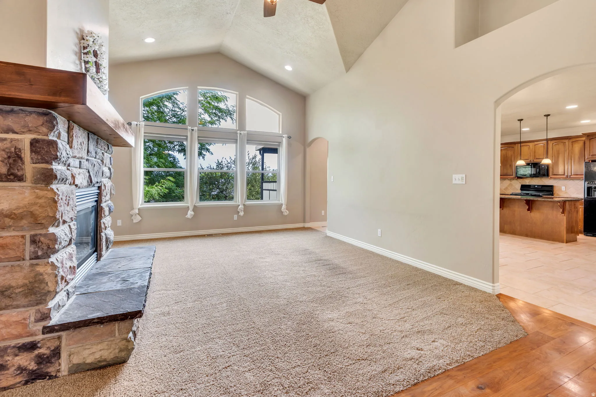 Vaulted ceiling with recessed lighting, stone-clad fireplace with wood mantle, large windows, and an arched doorway to the kitchen with wood-finish cabinetry
