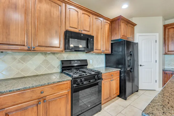 Wood cabinetry with dark hardware, stone-finish countertops, black appliances, tile backsplash, and tile flooring