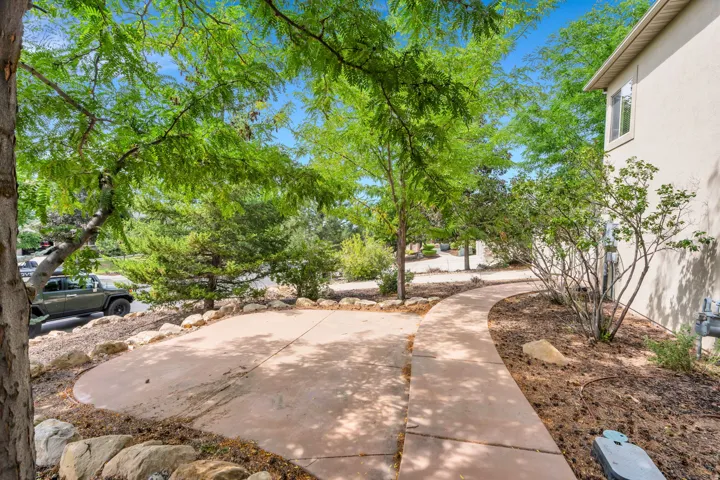 Curved concrete pathway, mature trees providing natural shade, concrete patio area, stucco exterior, and natural rock landscaping