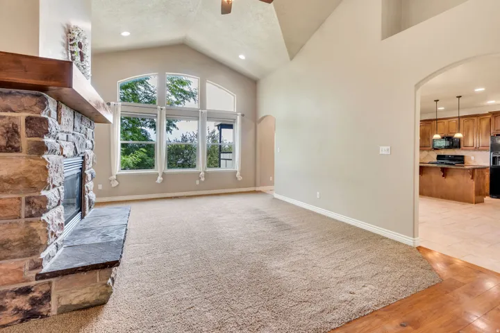 Vaulted ceiling with recessed lighting, stone-clad fireplace with wood mantle, large windows, and an arched doorway to the kitchen with wood-finish cabinetry