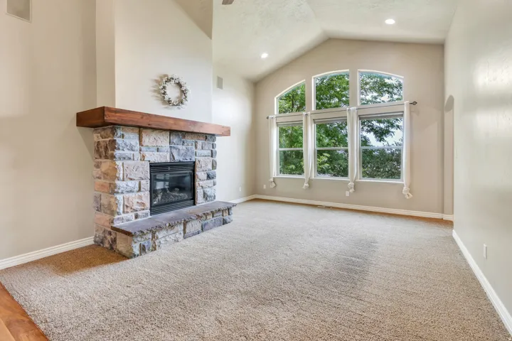 Vaulted ceiling with recessed lighting, stone-clad fireplace with wood mantel, large arched window, and neutral carpeting