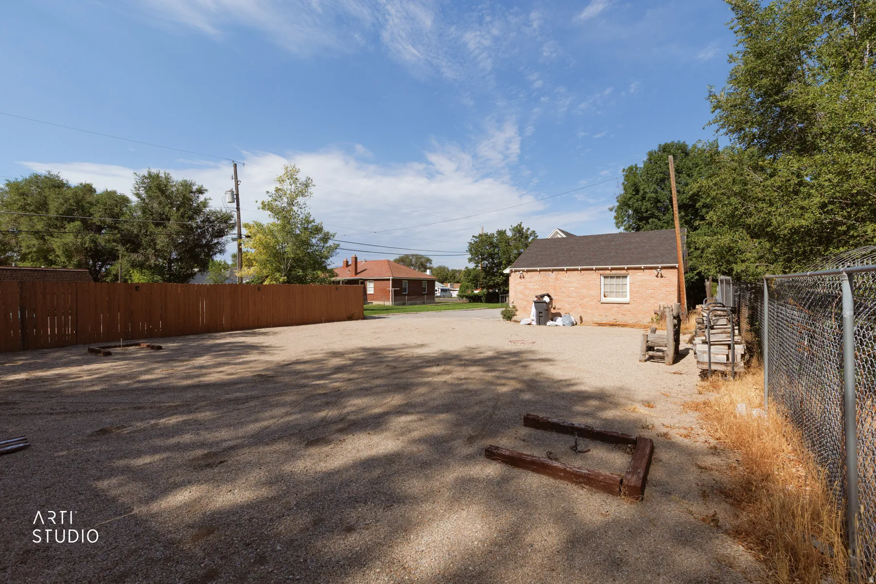 Gravel lot featuring a detached brick building with a shingled roof, a long wooden fence, a chain-link fence, and mature trees