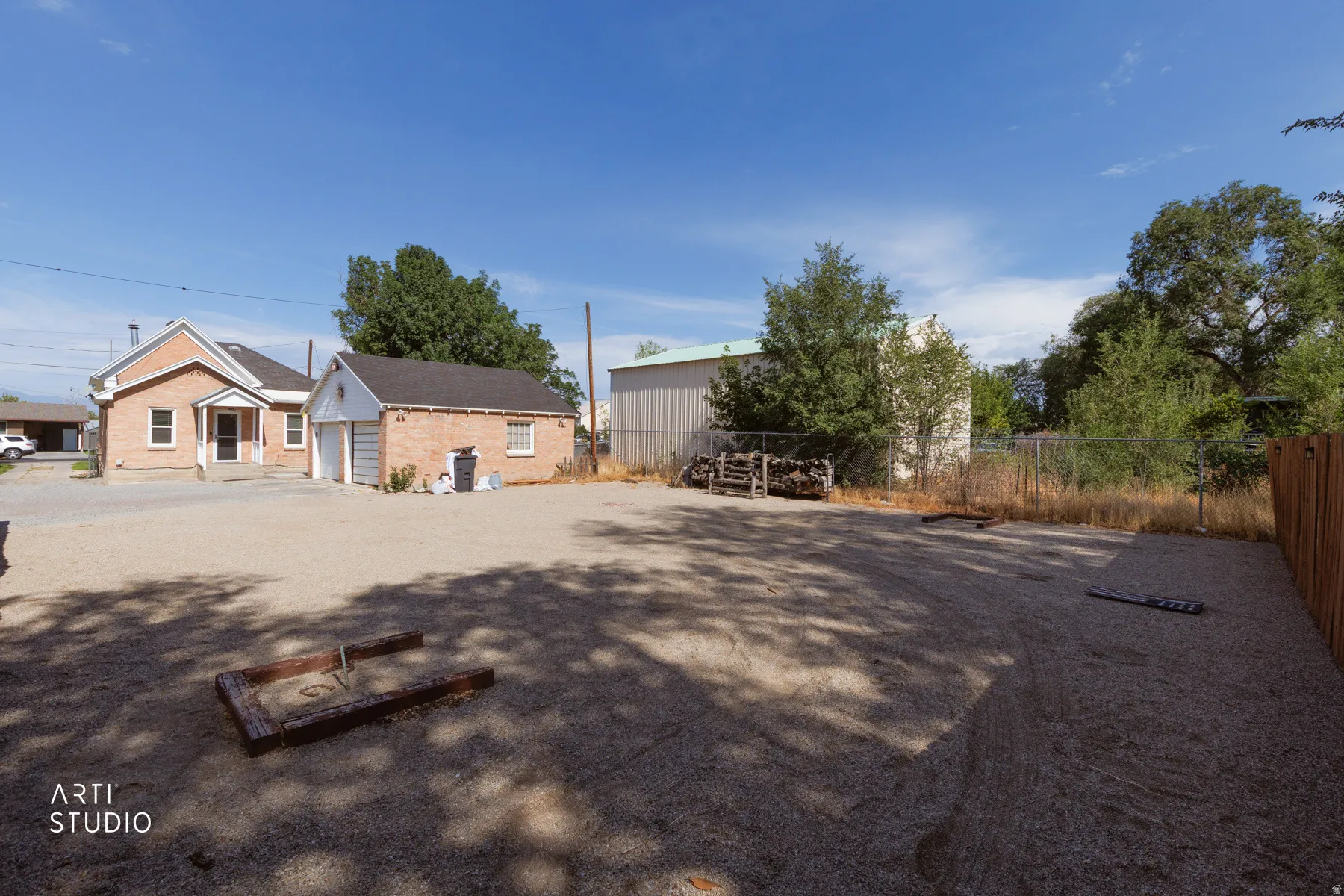 Expansive gravel lot featuring a detached brick house with an inviting porch, a separate brick garage, and mature trees providing shade