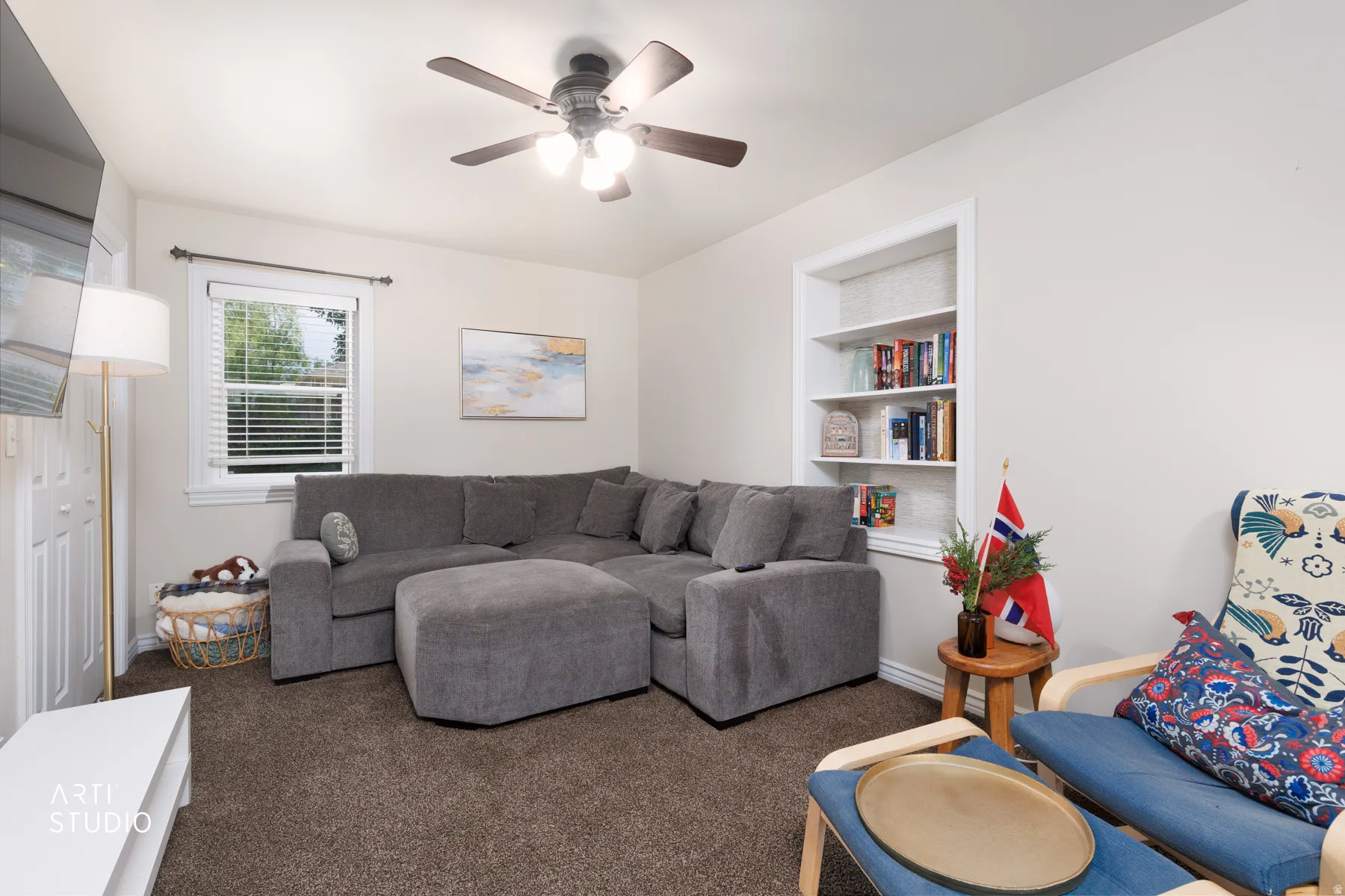 Carpeted living space featuring an integrated ceiling fan with lighting, a window with horizontal blinds, and a built-in recessed bookshelf