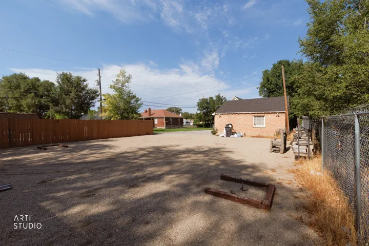 Gravel lot featuring a detached brick building with a shingled roof, a long wooden fence, a chain-link fence, and mature trees