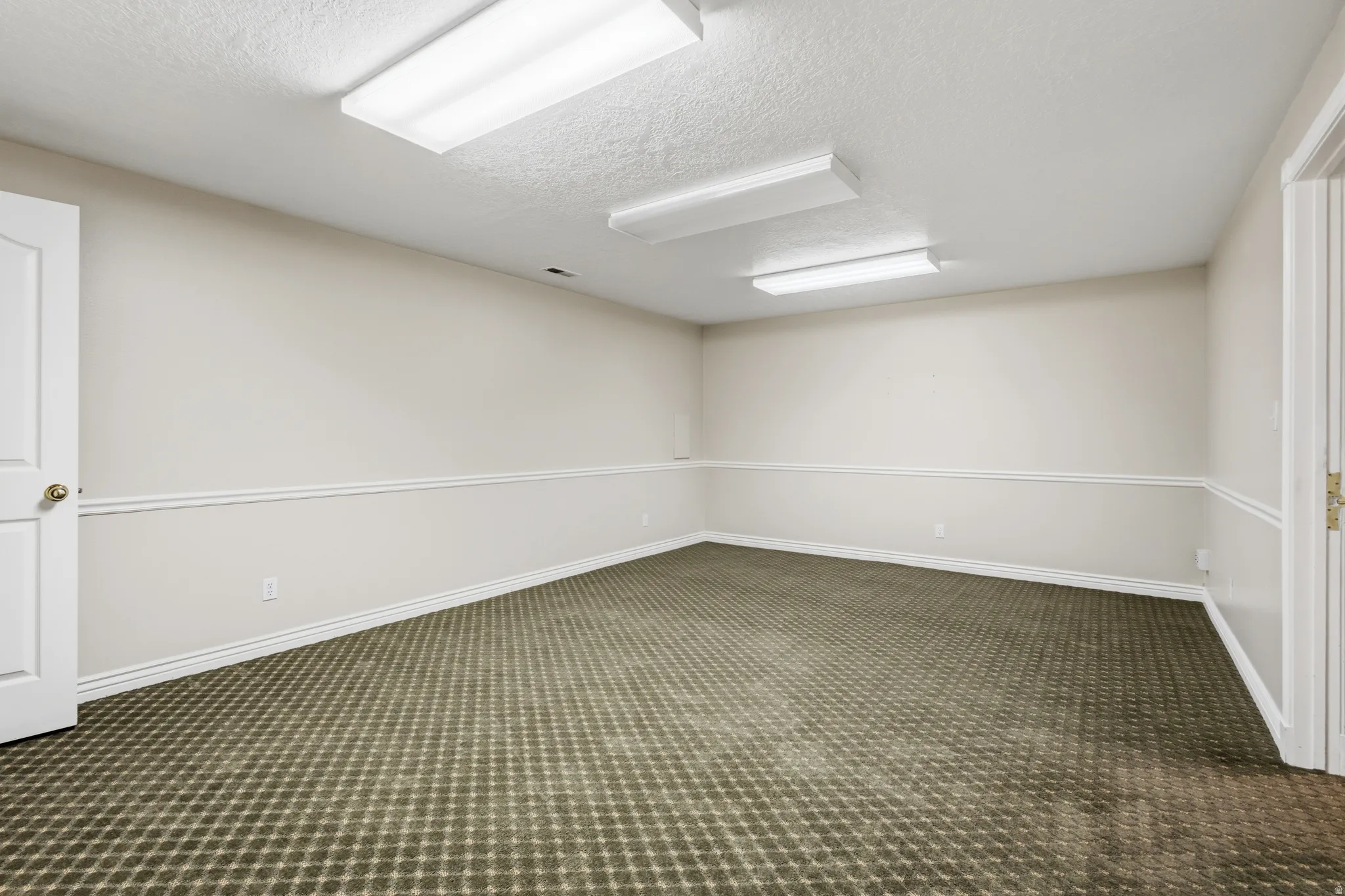 Spacious room featuring patterned carpet, light-toned walls with chair rail molding, and three rectangular ceiling light fixtures