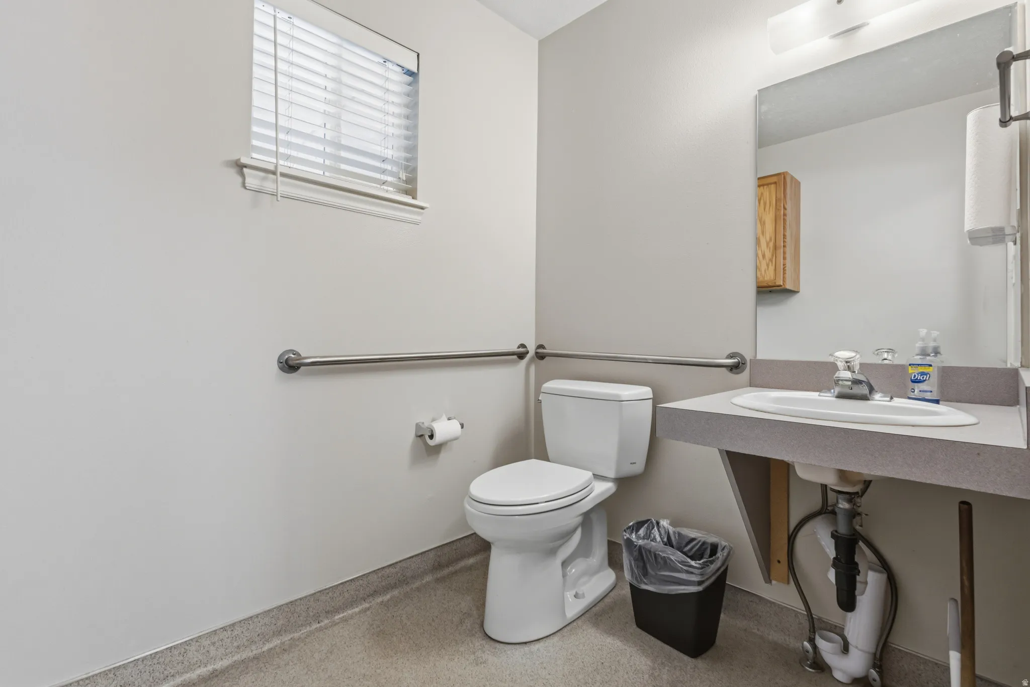 Bathroom featuring a window with blinds, a toilet, an accessible grab bar, a vanity with an integrated sink, and an overhead light fixture