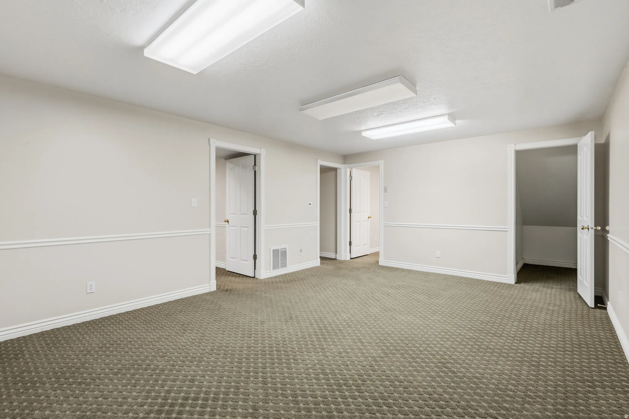 Spacious room featuring patterned carpeting, white wall paneling, multiple doorways, and fluorescent overhead lighting
