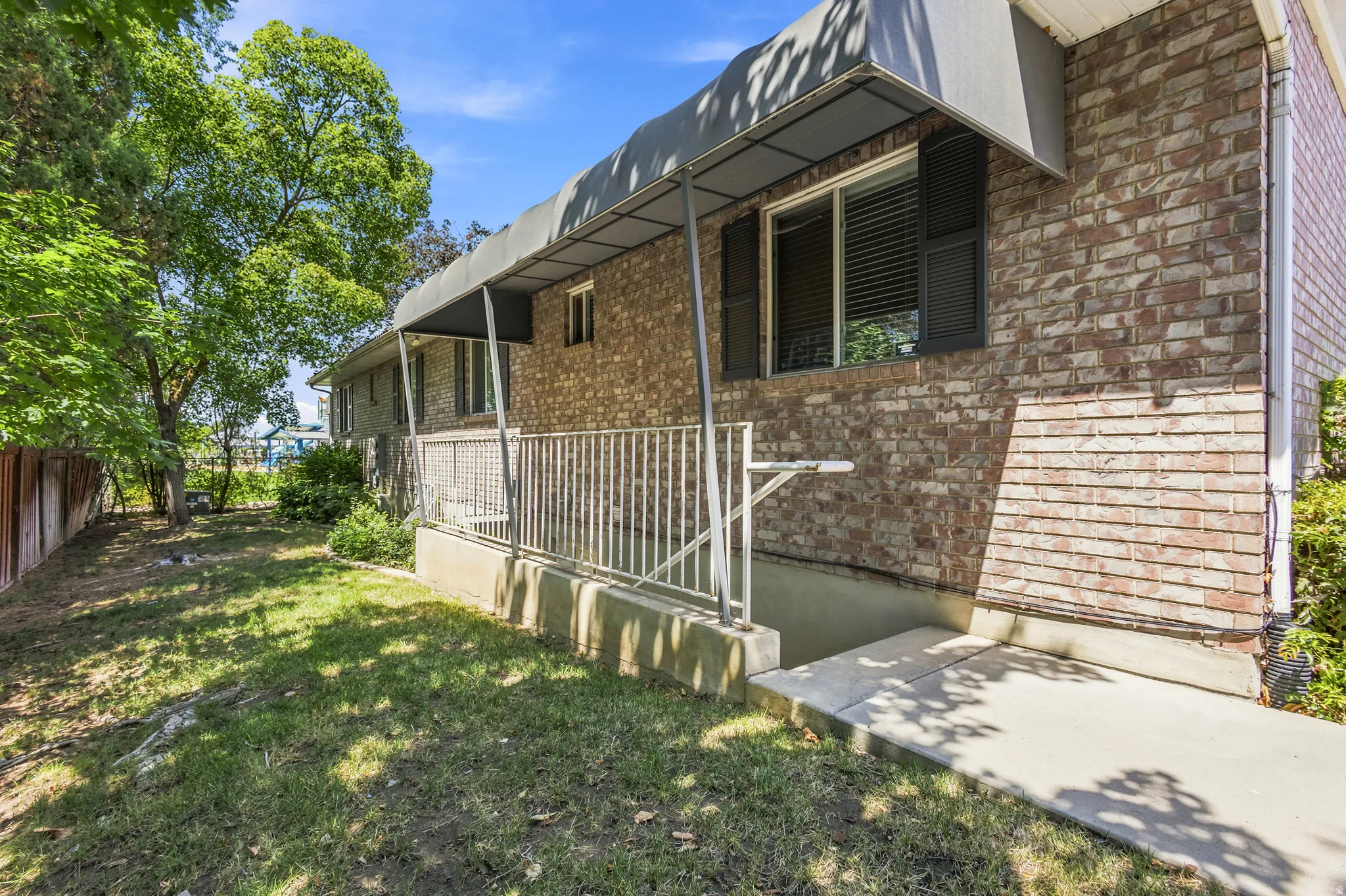 Brick exterior featuring multiple windows with black shutters, a covered porch with metal railing, and a mature tree