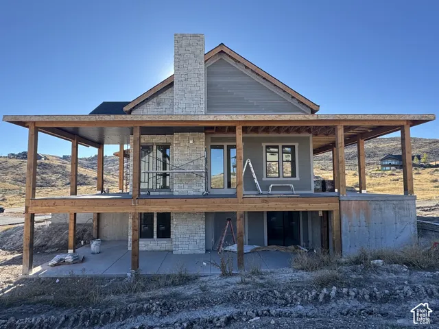 Exterior featuring horizontal siding, a prominent stone chimney, exposed timber post-and-beam construction, a covered upper-level deck, and a lower-level concrete patio