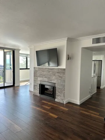 Living area featuring wood-finish flooring, a contemporary fireplace with stacked stone surround, crown molding, and a partial view of the kitchen with stainless steel appliances