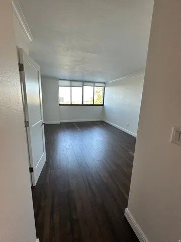 Spacious room featuring dark wood-finish flooring, white baseboards, crown molding, and large windows with horizontal blinds