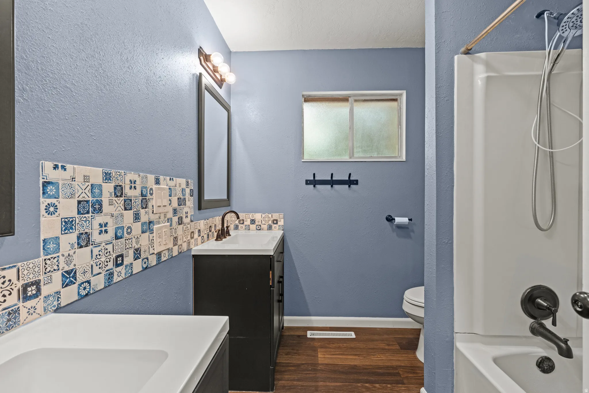 Bathroom featuring a white bathtub and shower combination, dark bronze fixtures, patterned tile accents, a dark wood-finish vanity and wood-finish flooring