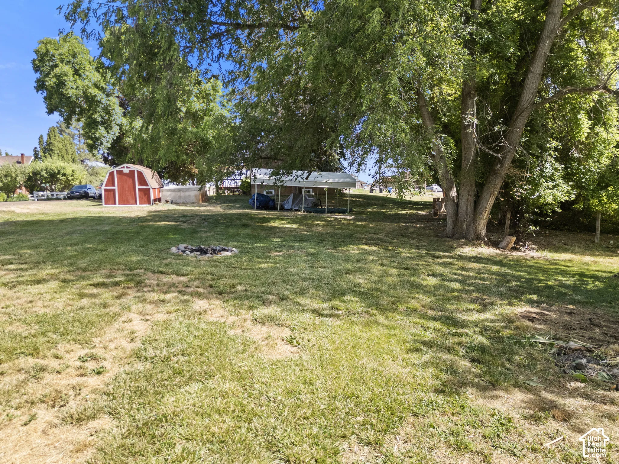 Expansive grassy yard featuring a red barn-style shed, mature shade trees, and a ground-level fire pit