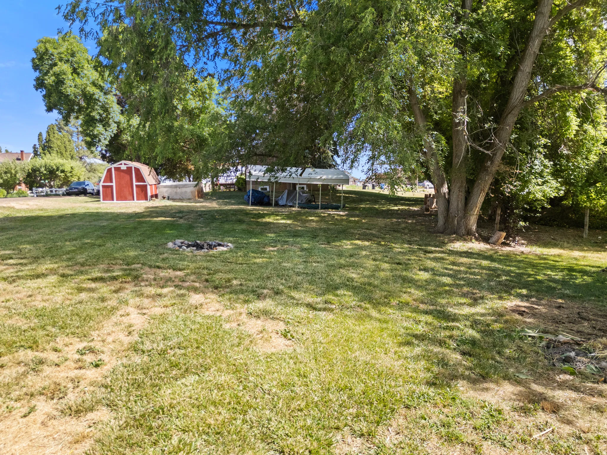 Expansive grassy yard featuring a red barn-style shed, mature shade trees, and a ground-level fire pit