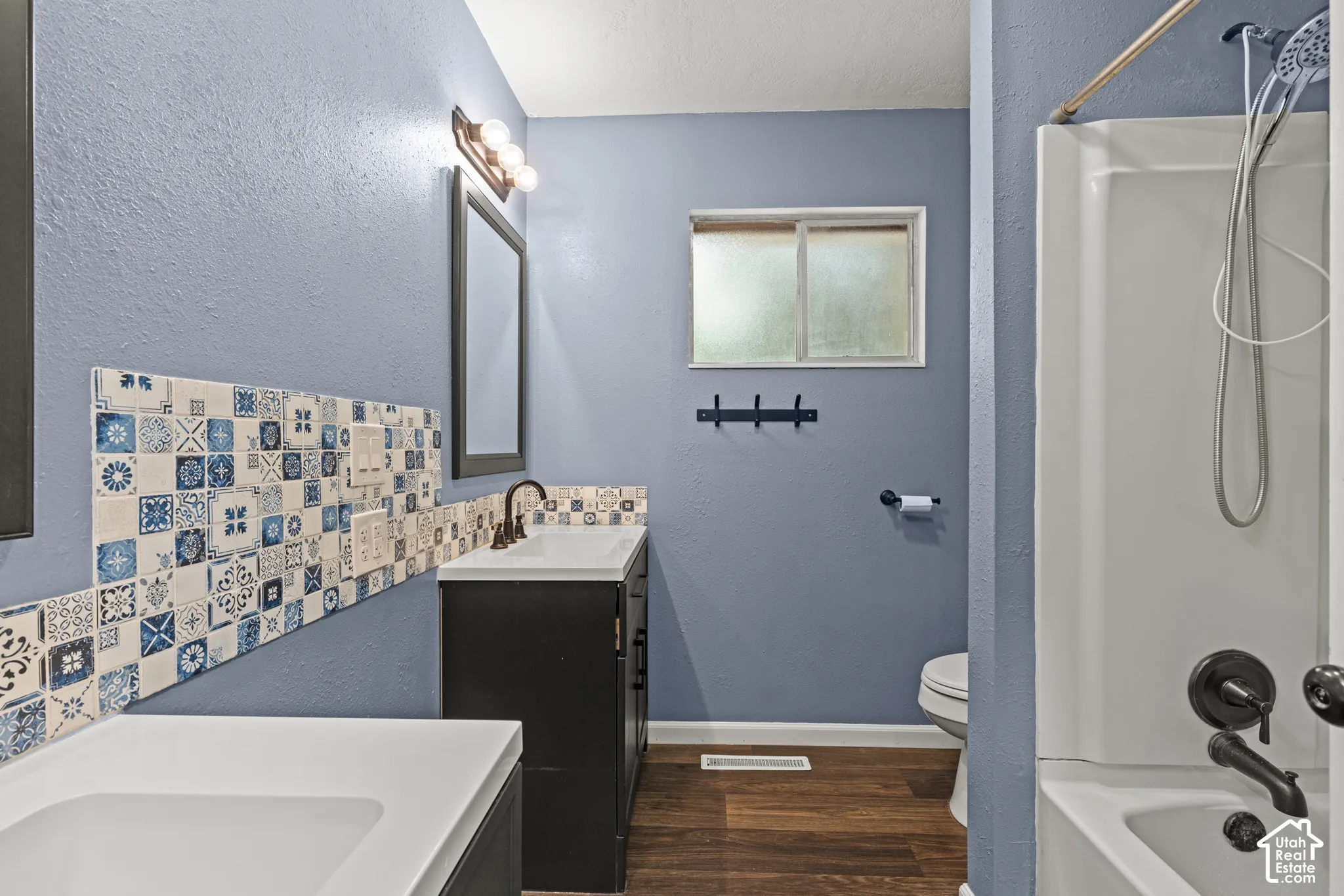 Bathroom featuring a white bathtub and shower combination, dark bronze fixtures, patterned tile accents, a dark wood-finish vanity and wood-finish flooring