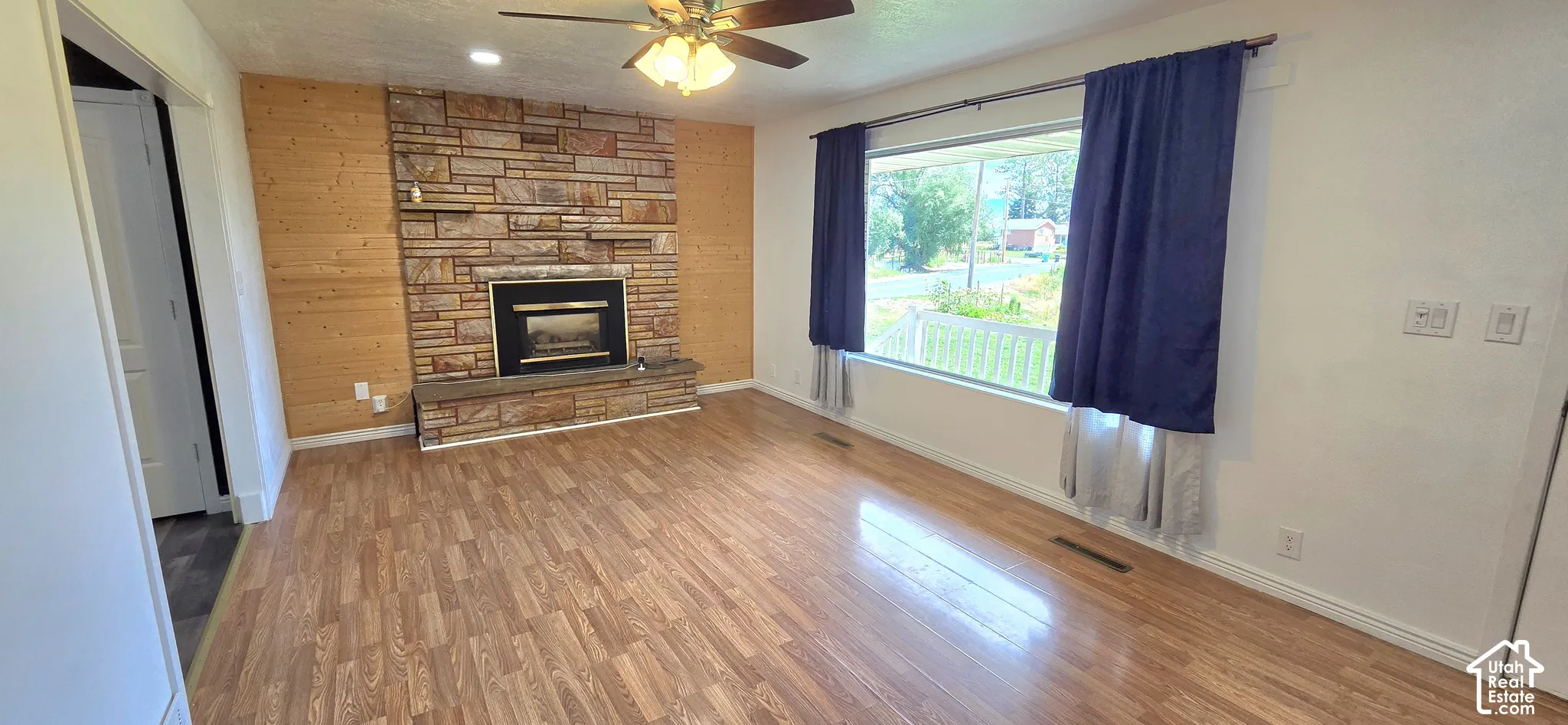 Wood-finish flooring, stone-clad fireplace, wood accent wall, ceiling fan with integrated lighting, and a large window