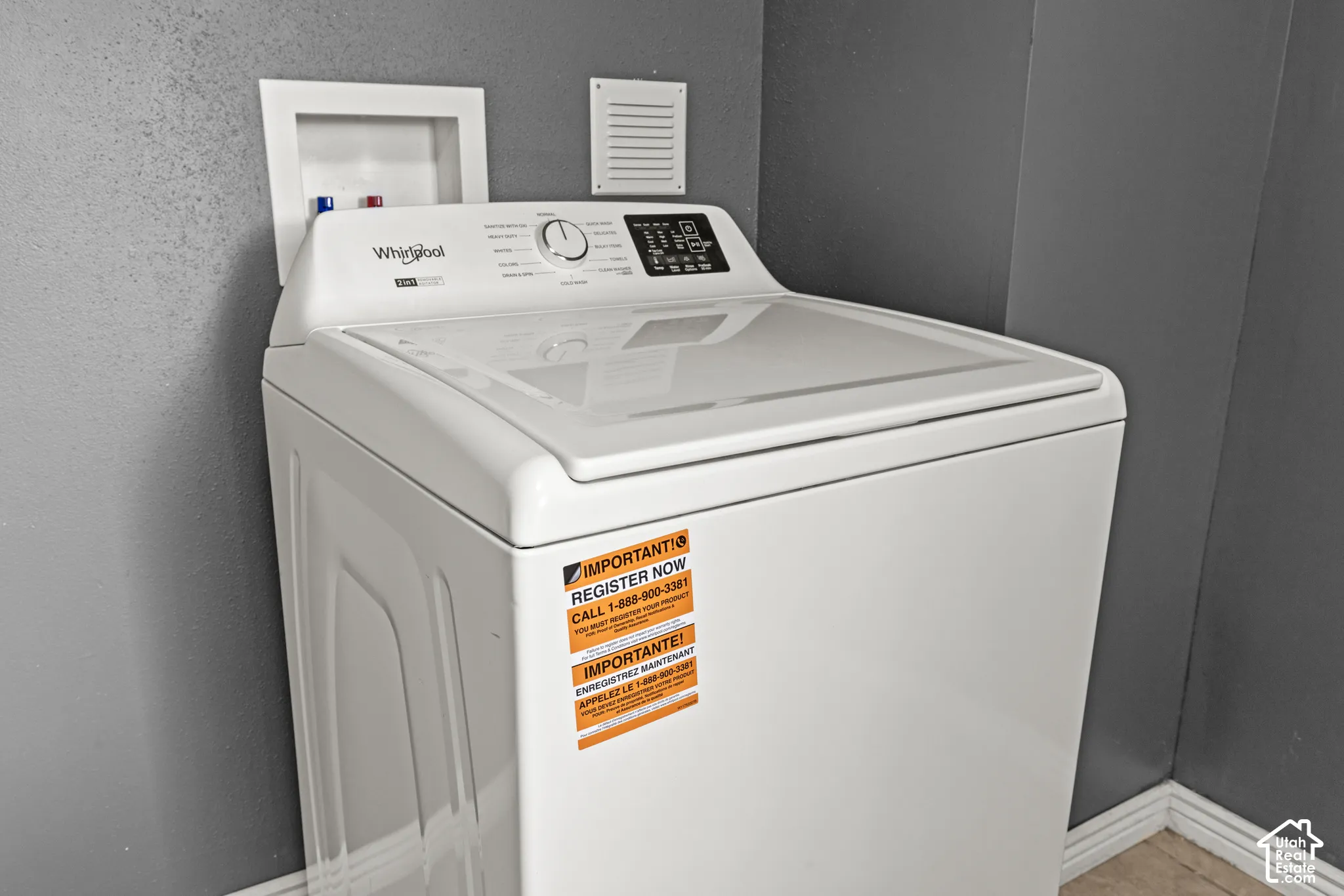 White top-loading washing machine with digital controls, featuring a water hookup box and an exhaust vent