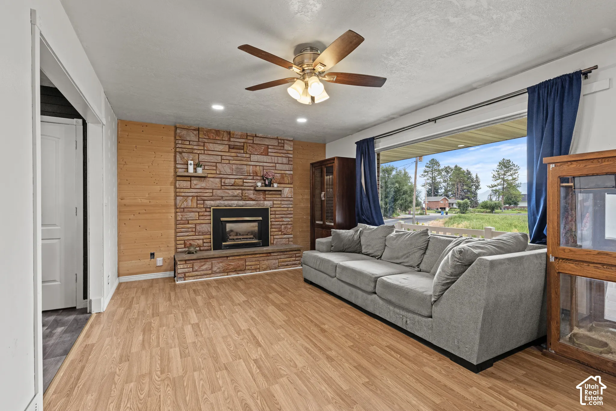 Spacious living area featuring a stone-clad fireplace with hearth, wood-finish flooring, a large window, recessed lighting, and a ceiling fan