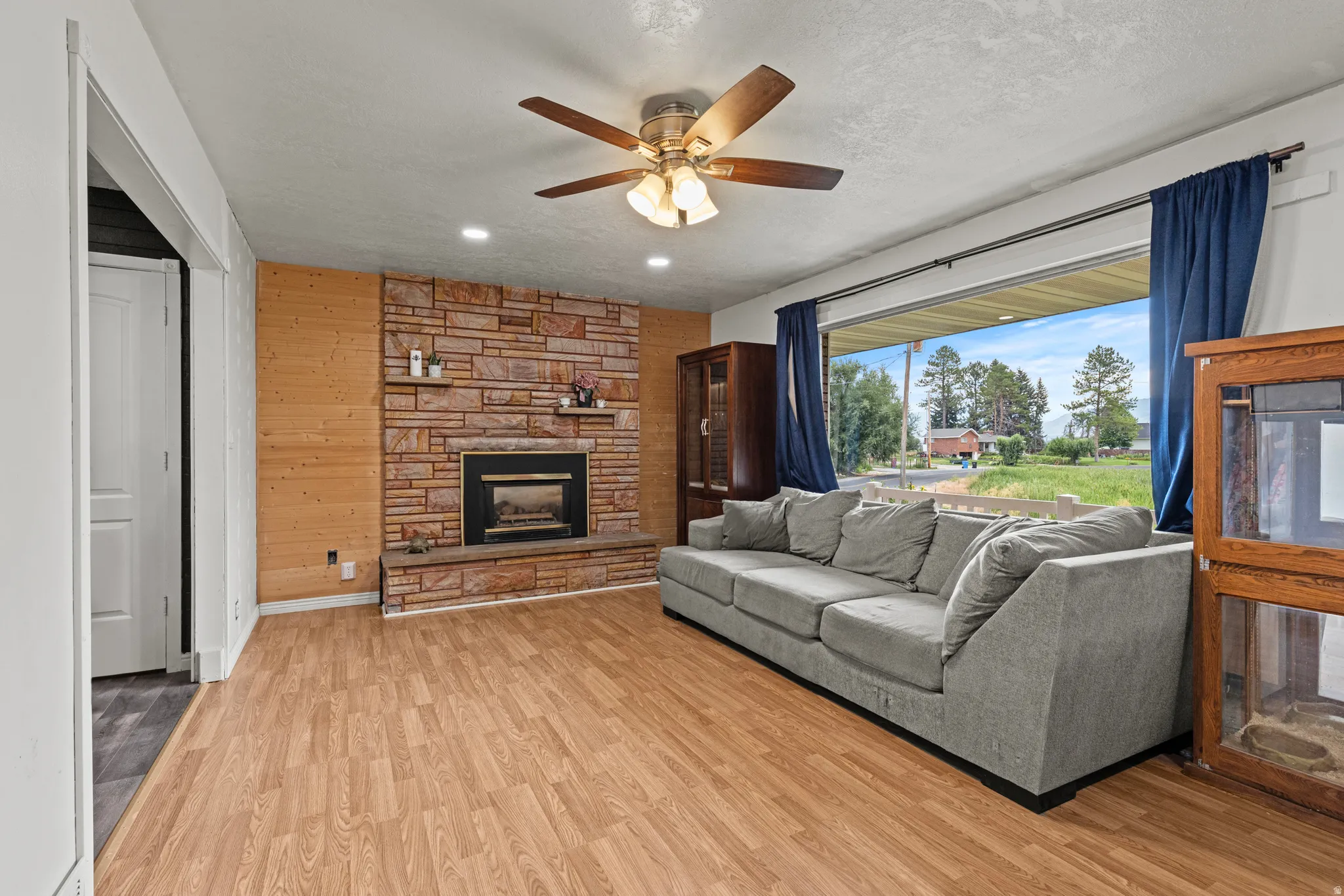 Spacious living area featuring a stone-clad fireplace with hearth, wood-finish flooring, a large window, recessed lighting, and a ceiling fan