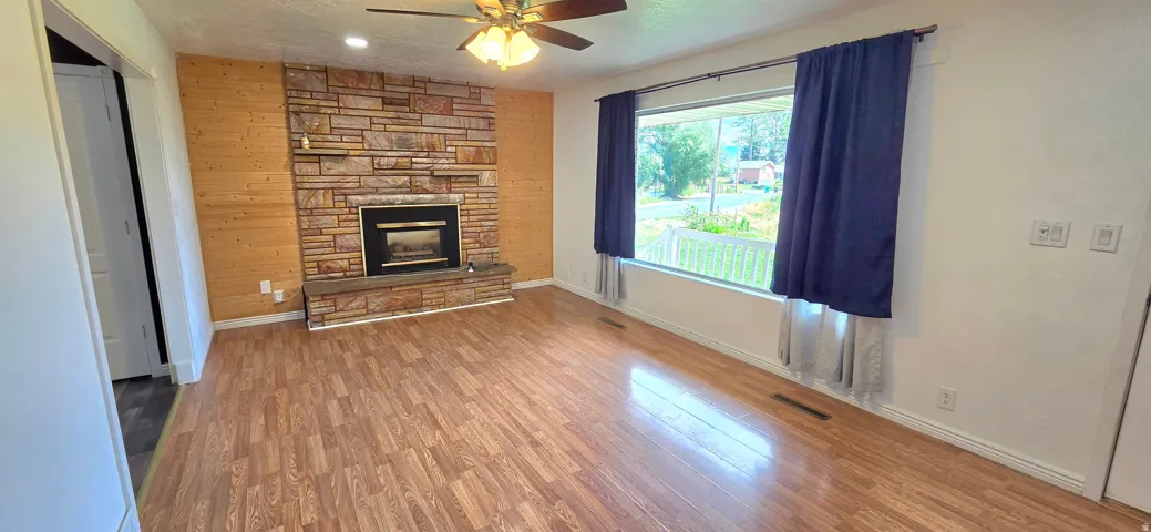 Wood-finish flooring, stone-clad fireplace, wood accent wall, ceiling fan with integrated lighting, and a large window