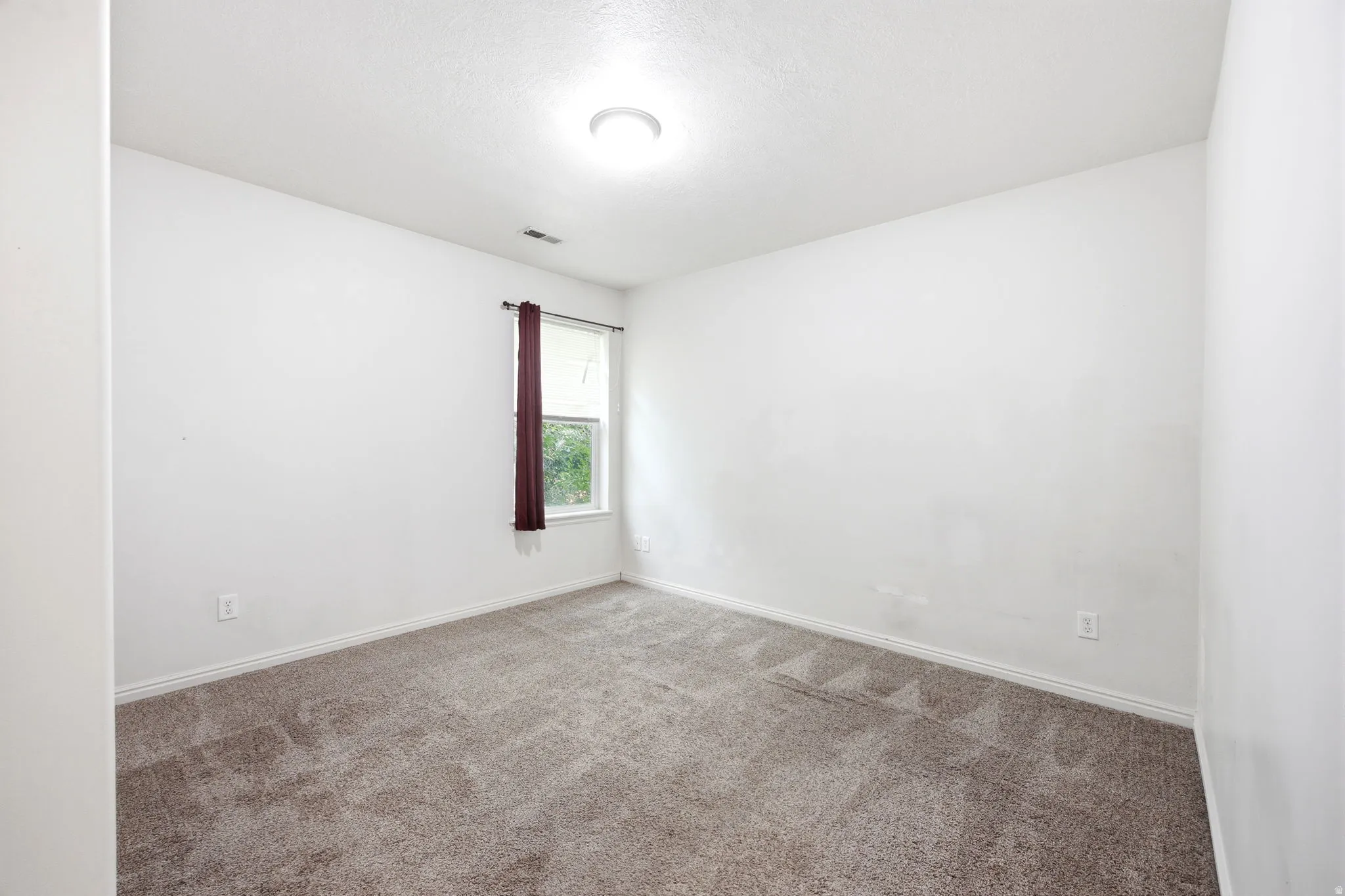 Carpeted interior space featuring white walls and a single window with dark red drapery
