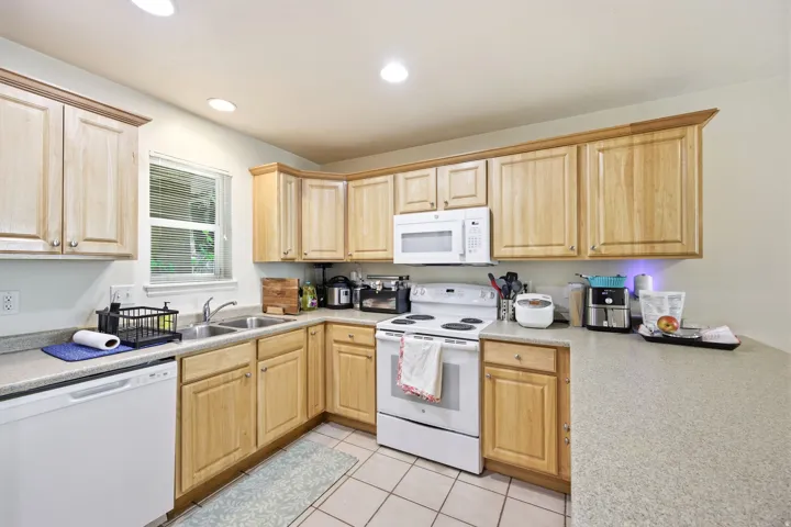 Kitchen featuring light wood cabinetry, recessed lighting, double basin stainless steel sink, light-toned countertops, and white appliances