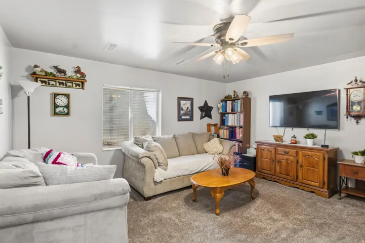 Spacious living area featuring neutral carpeting, light-toned walls, a ceiling fan with integrated lighting, and a horizontal window with blinds