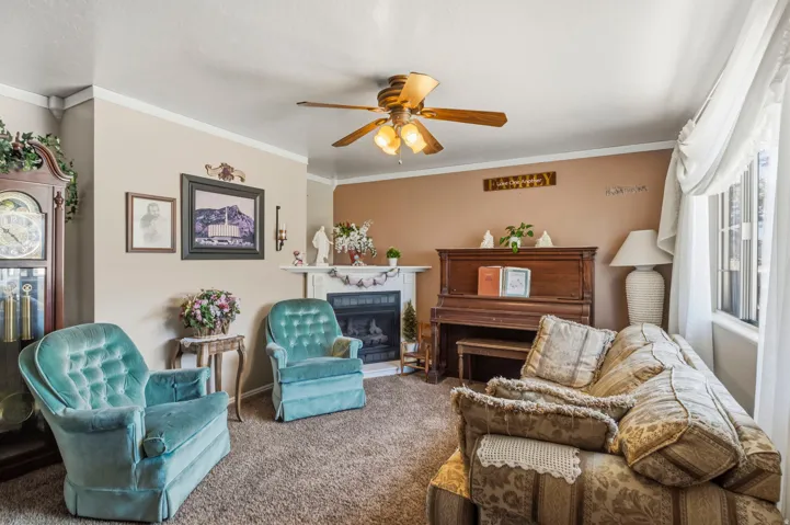 Living area with crown molding, ceiling fan with light fixture, central fireplace with mantle, and large window with transom