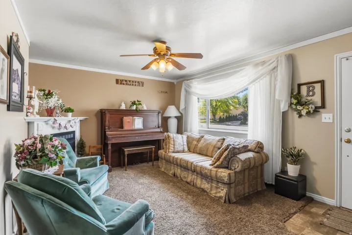 Carpeted living area featuring a decorative fireplace with a white mantel, a ceiling fan with integrated lighting, and a bay window with sheer curtains