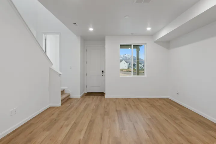 Spacious room featuring wood-finish flooring, white walls, recessed lighting, and a white entry door
