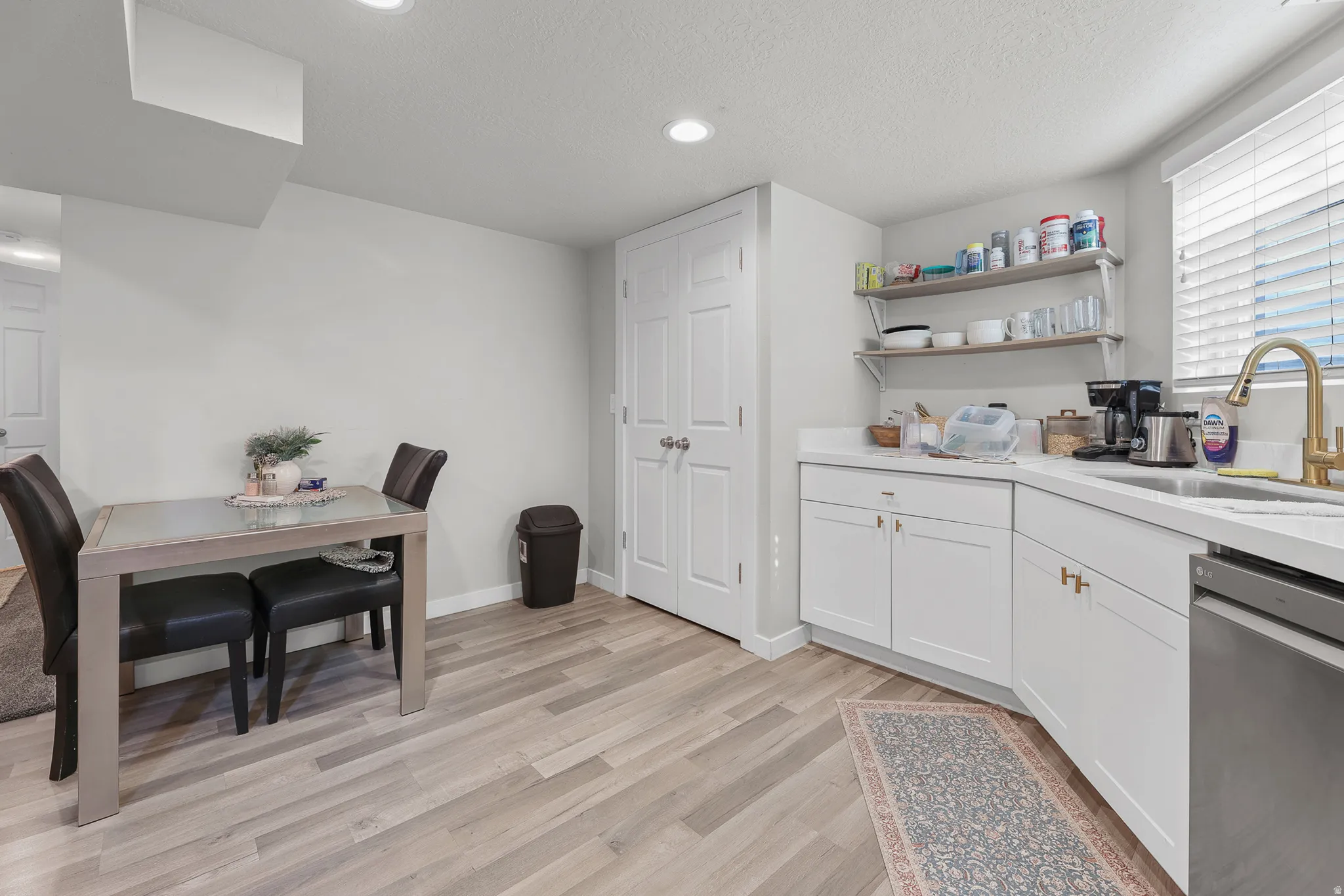 Kitchenette featuring white cabinetry, light-toned countertops, a stainless steel sink with a gold-finish faucet, and open shelving