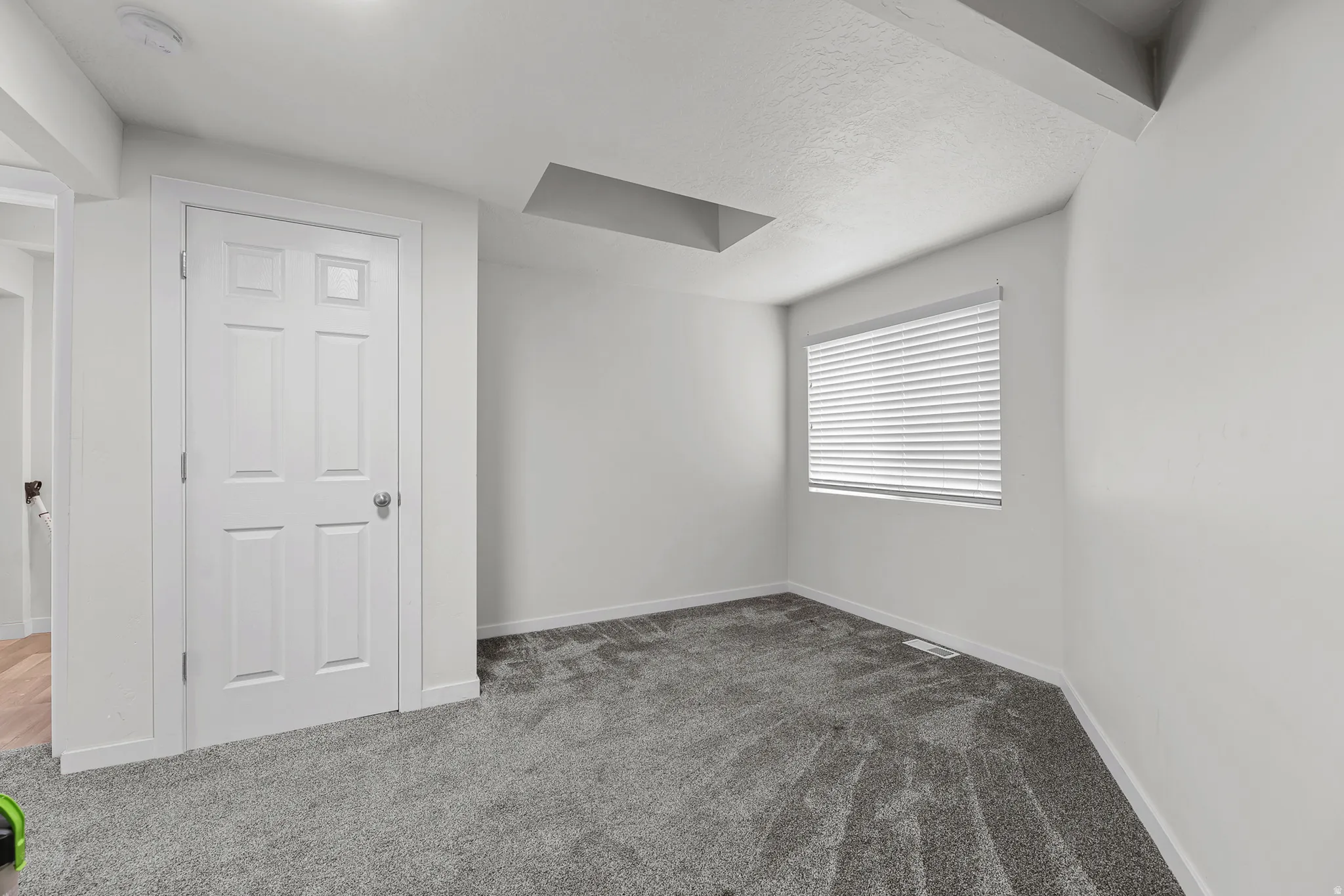 Room featuring plush grey carpet, a white paneled door, a window with horizontal blinds, white walls, and a white ceiling with visible structural beams