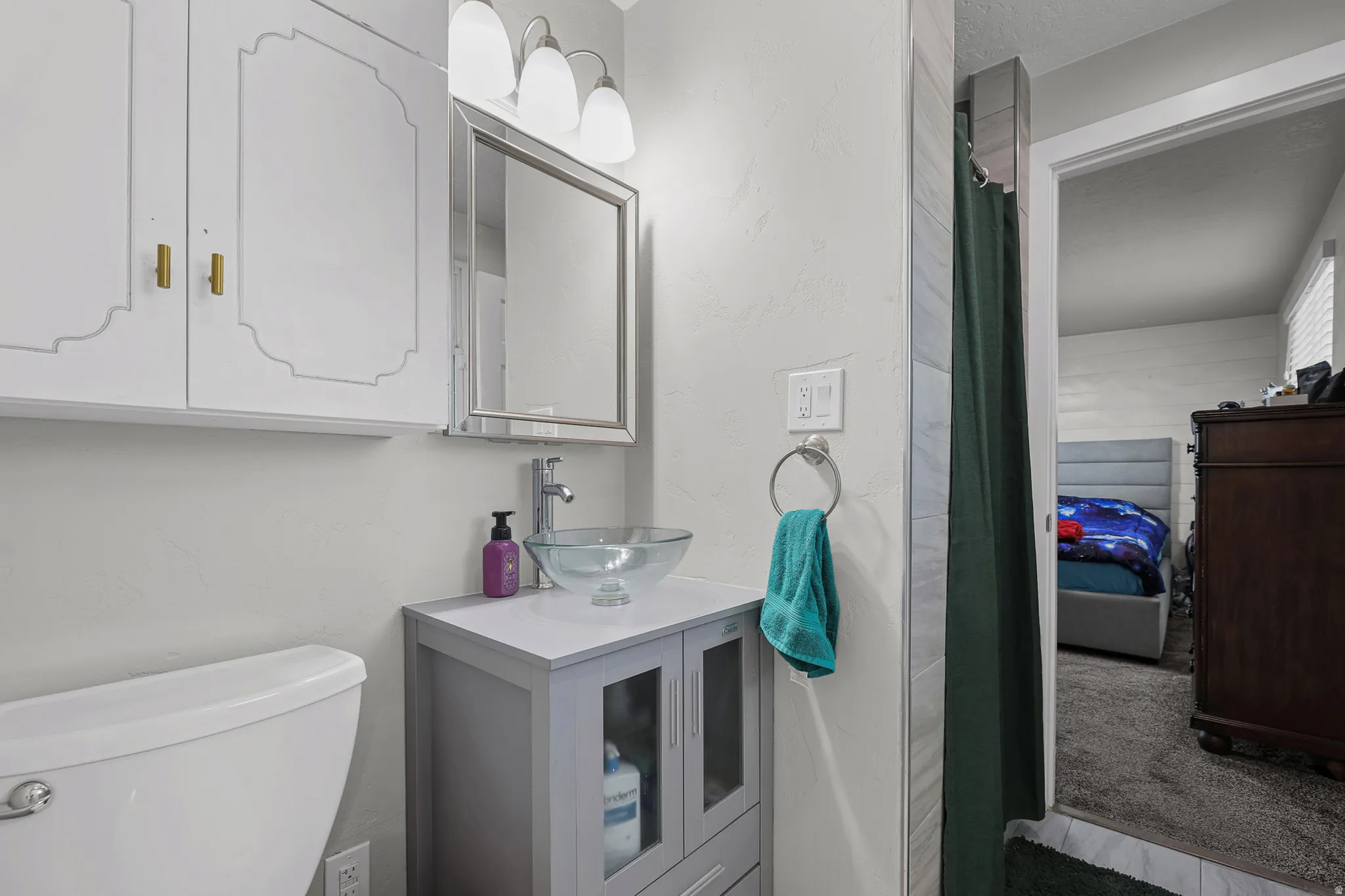 Bathroom featuring a vessel sink with a chrome faucet, light gray vanity with glass-front doors, white cabinetry with gold-toned hardware, and a framed mirror under a three-light fixture