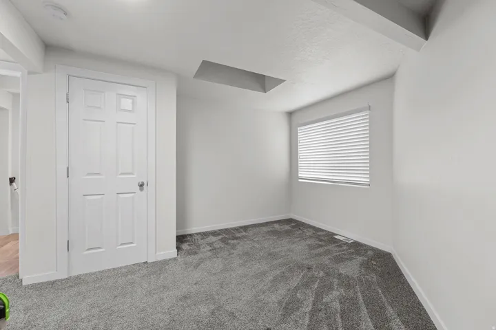 Room featuring plush grey carpet, a white paneled door, a window with horizontal blinds, white walls, and a white ceiling with visible structural beams