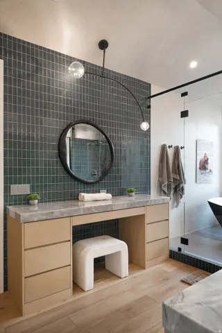 Light wood-finish flooring, built-in vanity with light wood-finish drawers, stone countertop, circular mirror, and dark tile wall