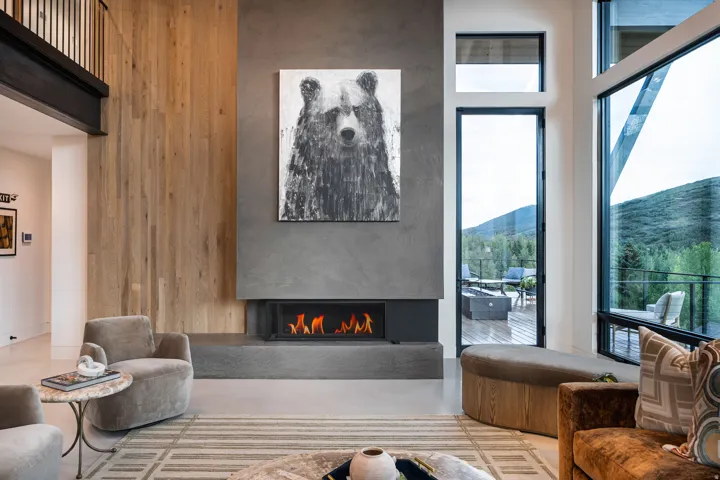 Integrated linear fireplace with a concrete surround, wood-paneled wall, large floor-to-ceiling windows, and a glass-paneled door leading to an outdoor deck