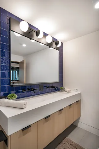 Dual integrated sinks with wall-mounted matte black faucets, light wood-finish cabinetry, and a full-width mirror with vanity lighting