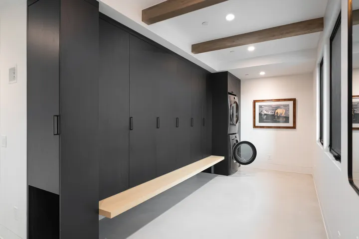 Integrated black cabinetry, light wood-finish floating bench, stacked washer and dryer, recessed lighting, and exposed ceiling beams