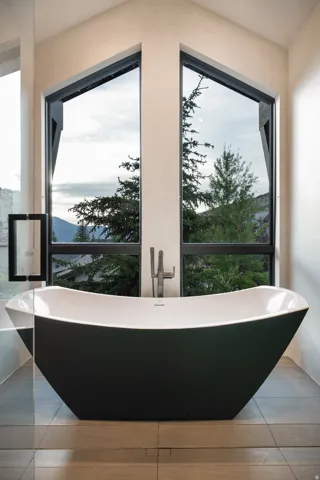 Freestanding soaking tub with a modern floor-mounted faucet, large windows with black frames, and tile flooring