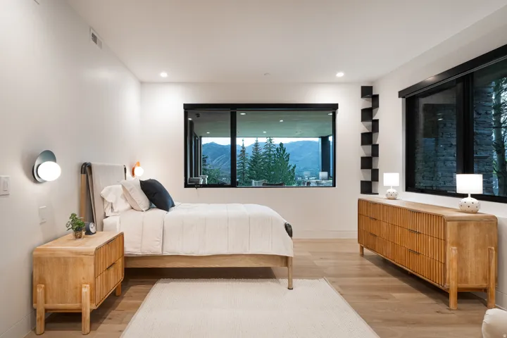 Hardwood flooring and recessed lighting illuminate a room with large windows, a built-in wall shelf, and modern wall sconces