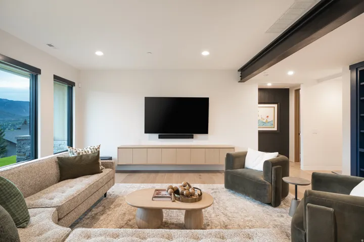 Spacious living area featuring large windows, wood-finish flooring, recessed lighting, a floating media cabinet, and an exposed structural beam