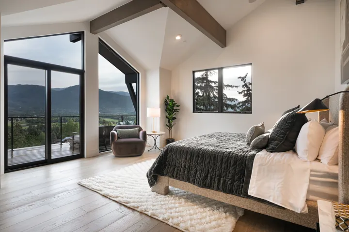 Angled ceilings with exposed wood beams, expansive floor-to-ceiling windows, direct balcony access, and wood-finish flooring