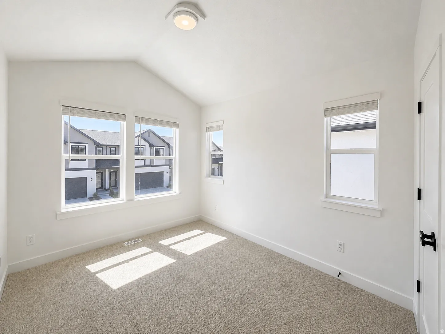 Vaulted ceiling room with neutral carpet flooring, three windows with blinds, a white paneled door with black hardware, and a flush-mount ceiling light fixture