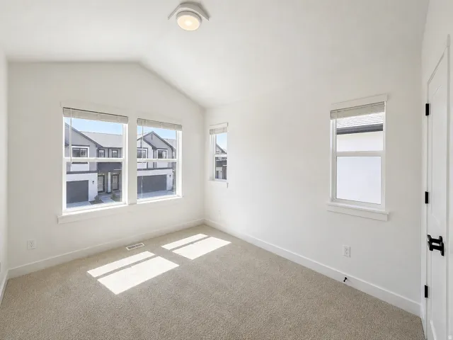 Vaulted ceiling room with neutral carpet flooring, three windows with blinds, a white paneled door with black hardware, and a flush-mount ceiling light fixture