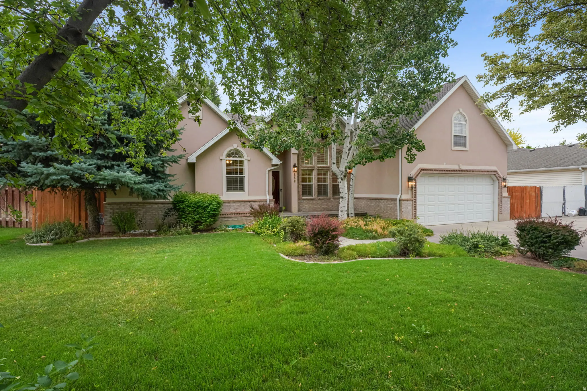 Stucco exterior residence featuring a two-car garage with a paneled door, arched window accents, and a mature treed lot