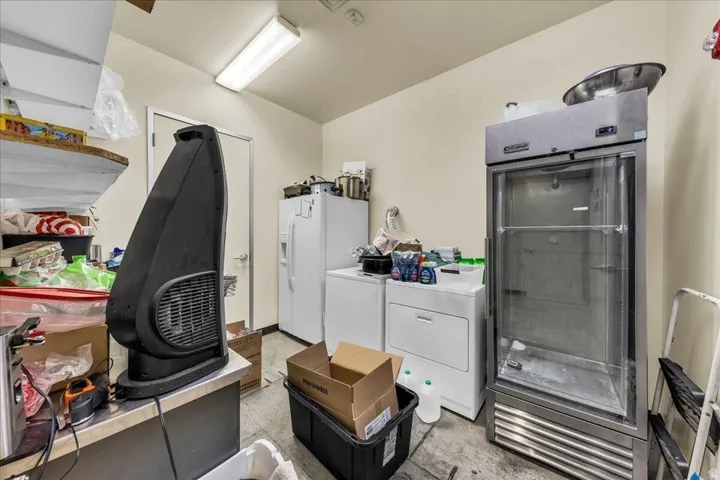 Utility space featuring a fluorescent ceiling light, a white refrigerator, a white washer, a white dryer, and a commercial-style glass door refrigerator